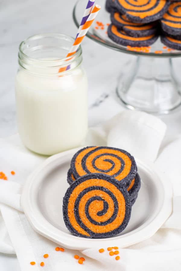 a pile of halloween pinwheel cookies on a white plate with a mason jar full of milk behind them. 
