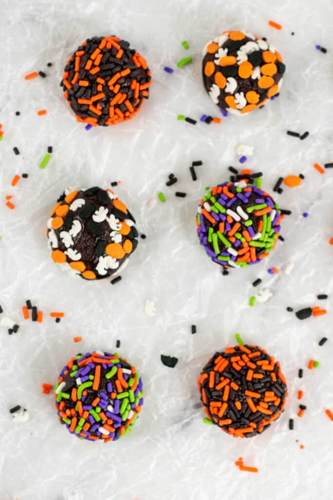 6 Halloween truffles lined up on a piece of parchment paper with sprinkles scattered around them. 
