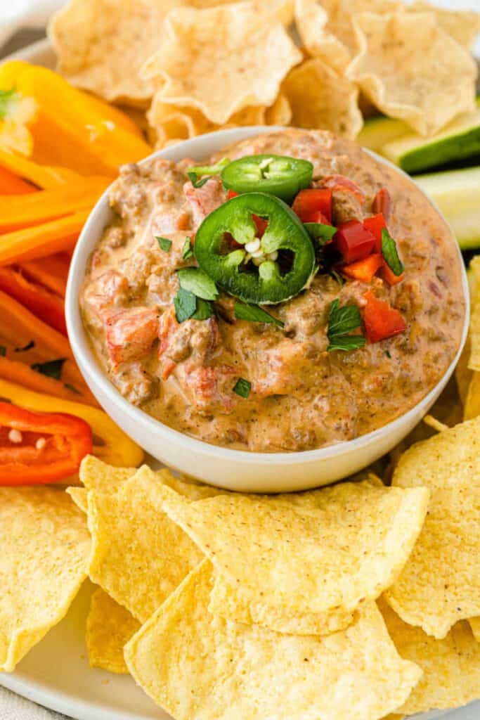 a bowl full of creamy sausage dip topped with jalapenos and red peppers surrounded by chopped vegetables and tortilla chips. 