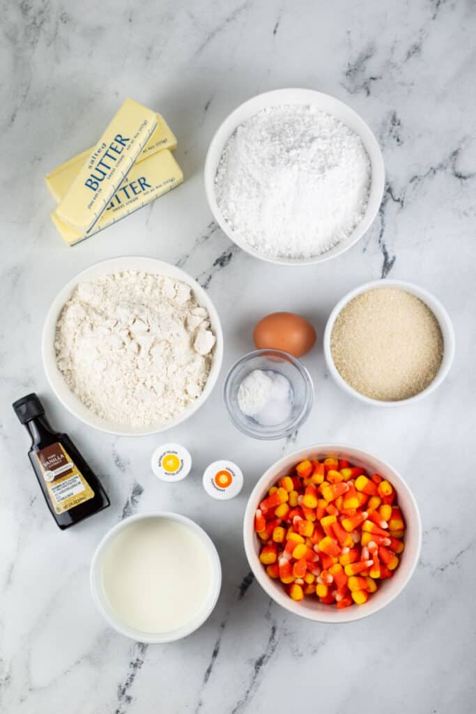 The ingredients for candy corn cookie bars on a white marble counter top. Flour, cream sugar, egg, vanilla, butter, powdered sugar, yellow and orange food coloring and candy corn. 