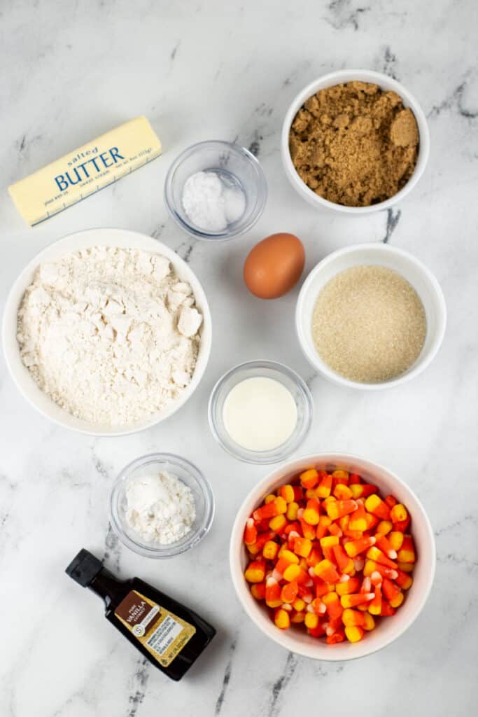 The ingredients for candy corn cookies on a white marble background. Butter, brown sugar, white sugar, egg, flour, heavy cream, salt, baking soda, vanilla and candy corn. 