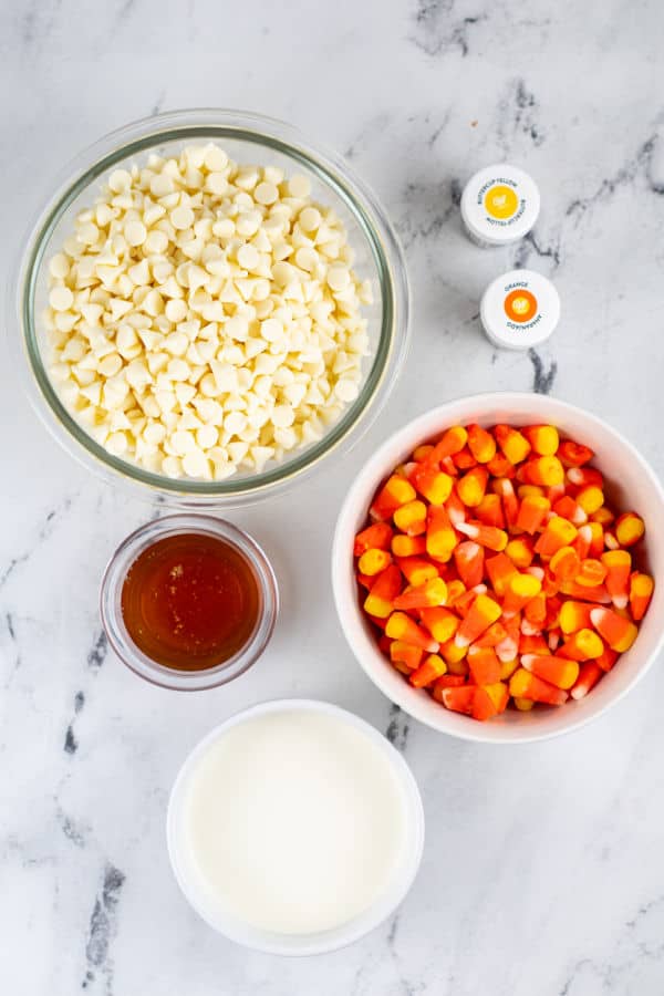 The ingredients for candy corn fudge on a white marble background. A bowl of white chocolate chips, honey, heavy cream, yellow and orange food coloring and candy corn. 