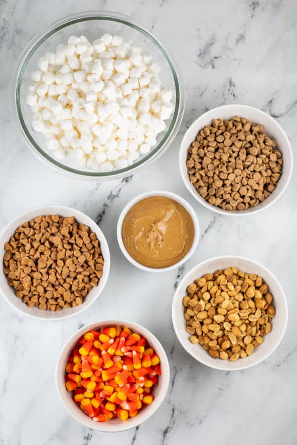 The ingredients for candy corn marshmallow bars on a marble counter top. Marshmallows, peanuts, peanut butter, peanut butter chips, butterscotch chips and candy corn. 