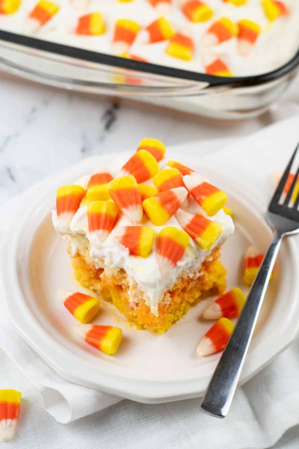 A piece of candy corn poke cake on a small white plate with a fork next to it. 