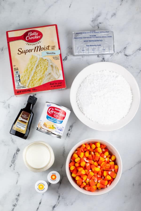 The ingredients for candy corn poke cake. A cake mix, cream cheese, powdered sugar, sweetened condensed milk, vanilla, cream, orange and yellow gel food coloring and candy corn. 