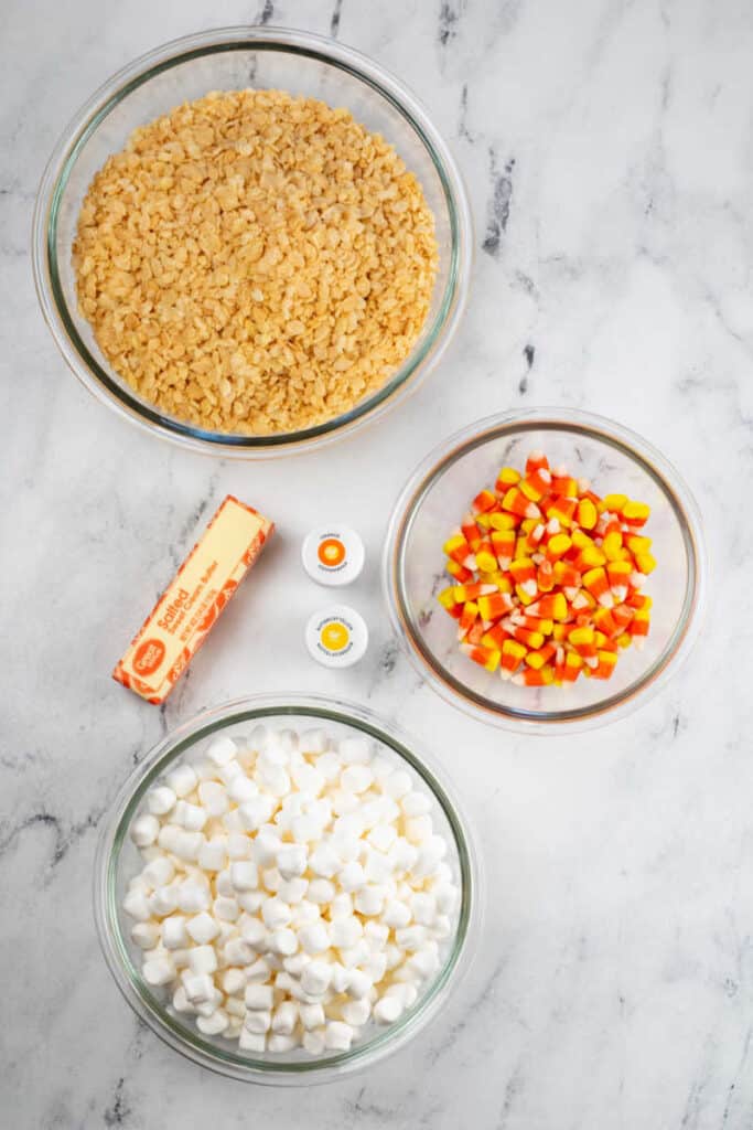 The ingredients for candy corn rice krispie treats on a marble countertop. A bowl full of rice cereal, butter, marshmallows, orange and yellow food coloring and candy corn. 