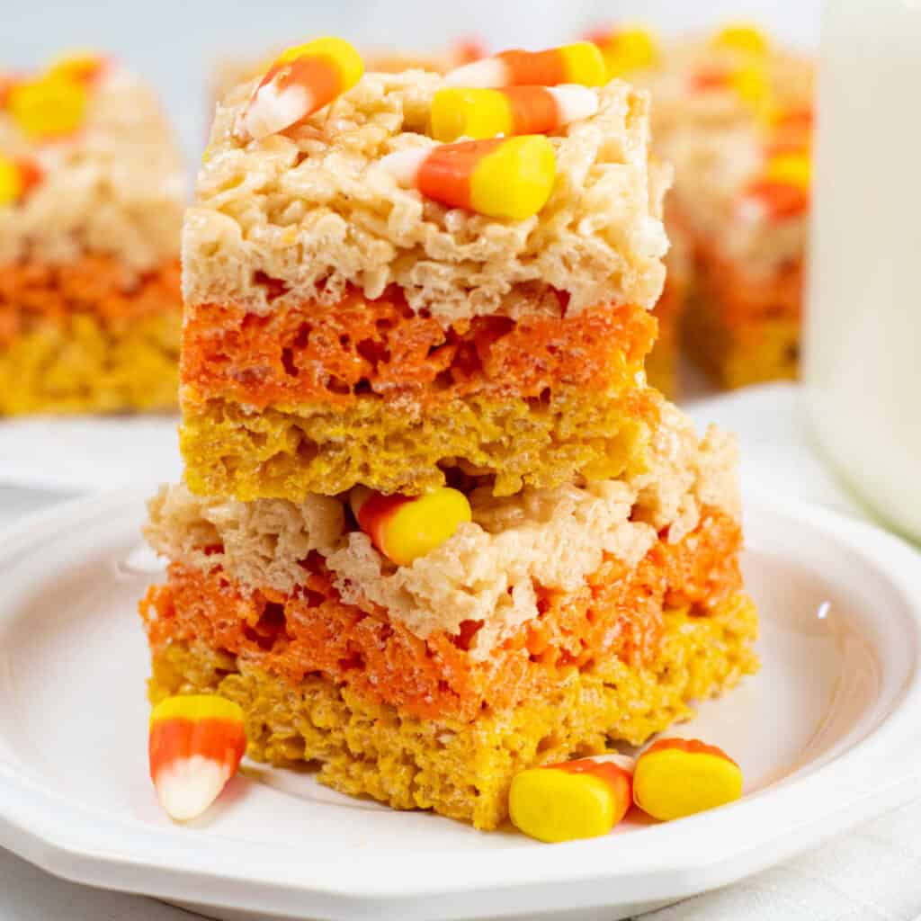 A stack of yellow, orange and white rice krispies treats topped with candy corn on a white plate. 