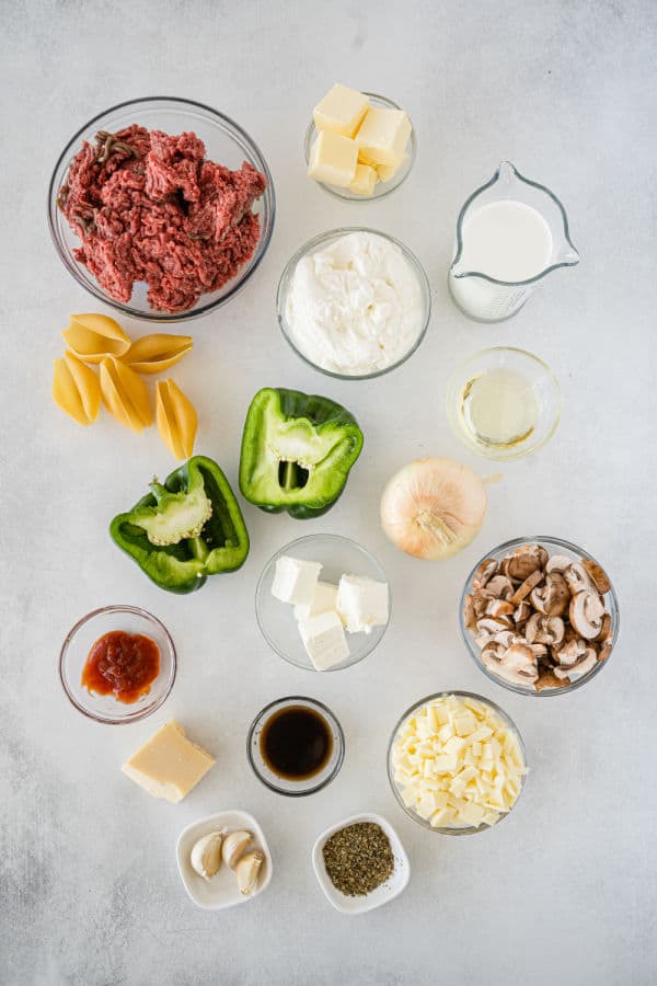 The ingredients for Philly cheesesteak stuffed shells on a white marble counter top. Ground Beef, butter, ricotta cheese, half and half, pasta shells, onion, olive oil, green peppers, mushrooms, cream cheese, ketchup, Worchestershire sauce, Garlic, salt, provolone cheese and parmasean cheese. 