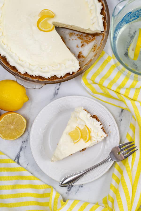 A slice of lemon pie on a white plate nest to it sits sliced lemons and the pie pan with a slice removed. 