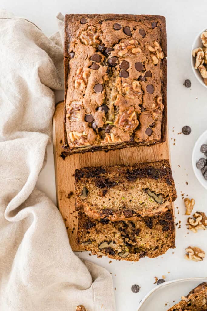 A loaf of chocolate chip zucchini bread partially sliced on a wooden chopping block. 