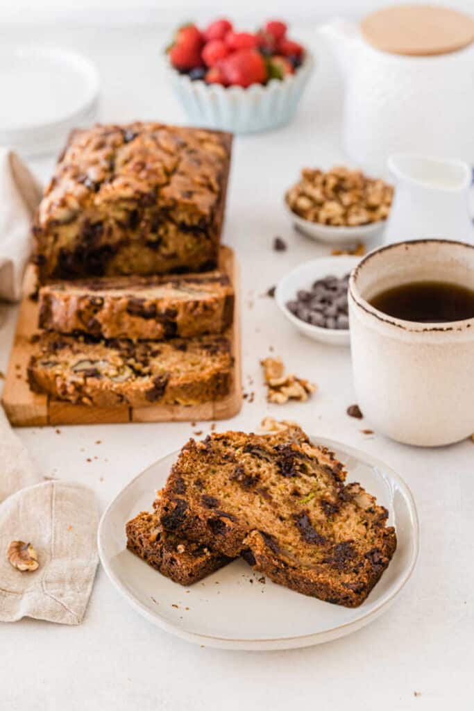 A table with a white plate with two slices of chocolate chip zucchini bread, a cup of coffee and a loaf of zucchini bread in the background. 