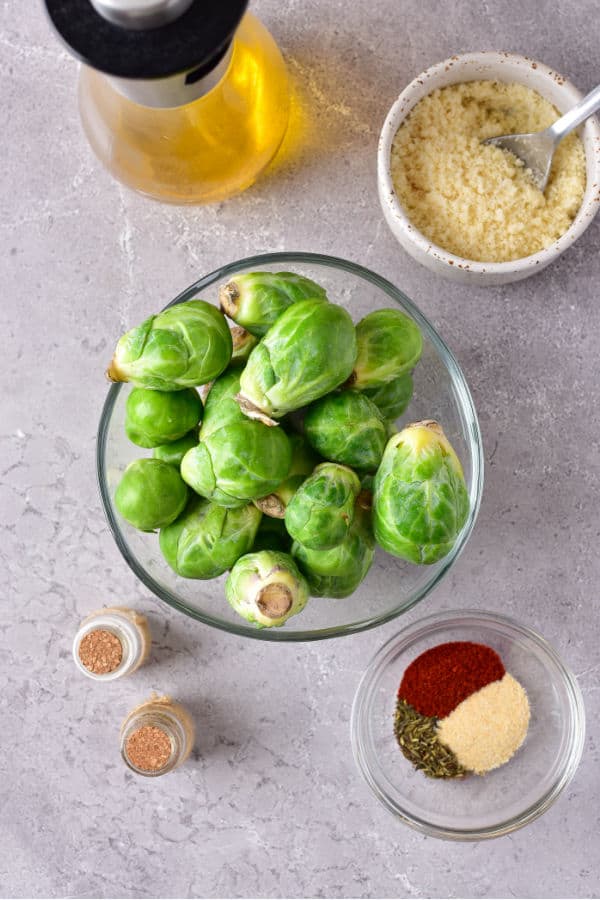 The ingredients for air fryer parmesan roasted brussels sprouts  on a grey marble counter. Brussels sprouts, salt, smoked paprika, garlic powder, thyme, oil and parmesan cheese. 