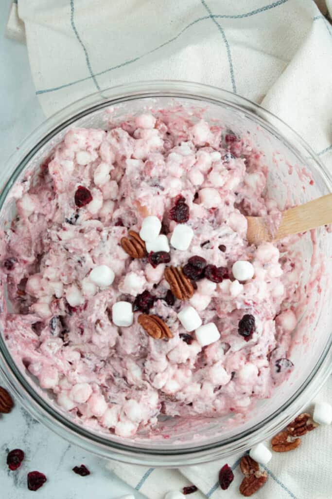 A clear glass bowl full of cranberry fluff salad topped with pecans, marshmallows and dried cranberries. 
