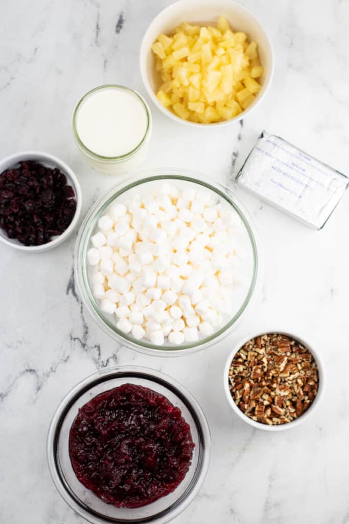 The ingredients for cranberry fluff on a white marble background. Marshmallows, cranberry sauce, dried cranberries, pineapple, cream cheese, and pecans 