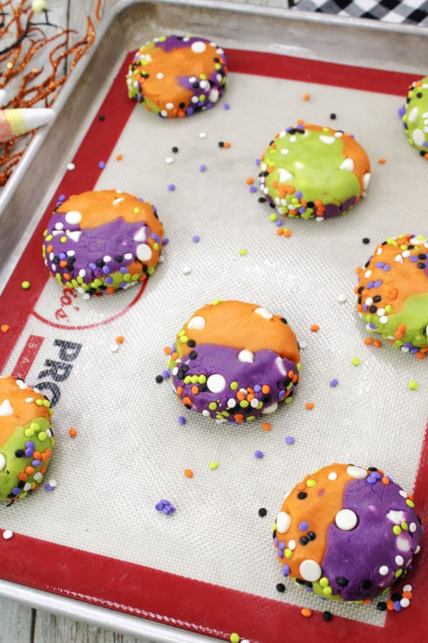 A lined baking sheet is topped with unbaked cookies. The cookies are orange purple and green with white chocolate chips and colorful sprinkles 