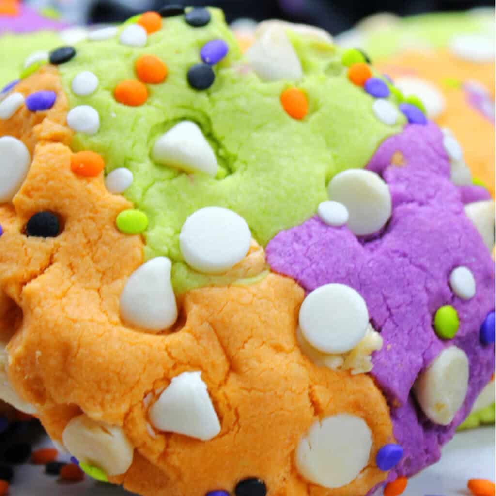A close up of a green, orange and purple cookie with white chocolate chips and colorful sprinkles 