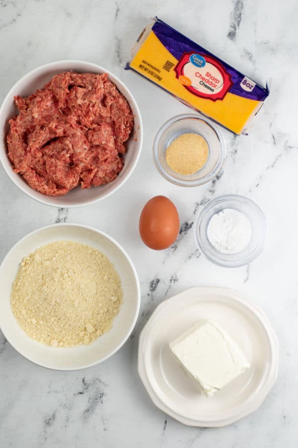 The ingredients for keto sausage balls on a marble background. Sausage, cheddar cheese, almond flour, egg, cream cheese, baking powder and onion powder. 