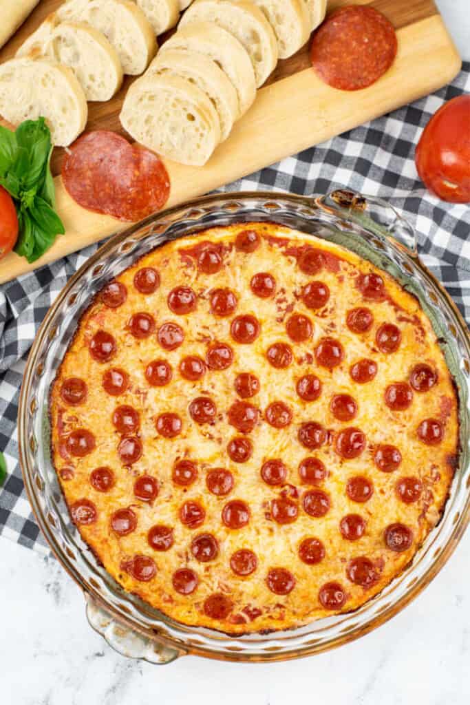 A pie pan full of warm pizza dip. Next to it sits a chopping block with slices of baguette on it. 