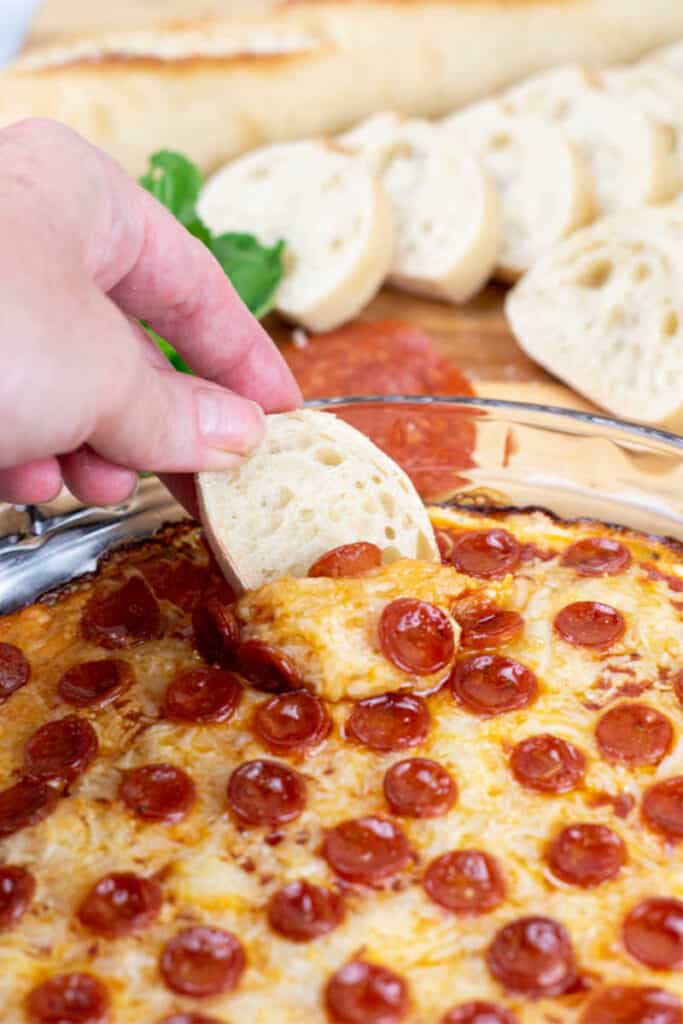 A hand dipping a slice of baguette into warm pizza dip. 