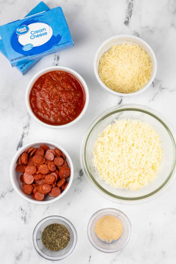Ingredients for pizza dip on a marble counter top. Cream cheese, shredded parmesan cheese, shredded mozzarella cheese, mini pepperoni, pizza sauce, garlic powder and Italian seasoning. 