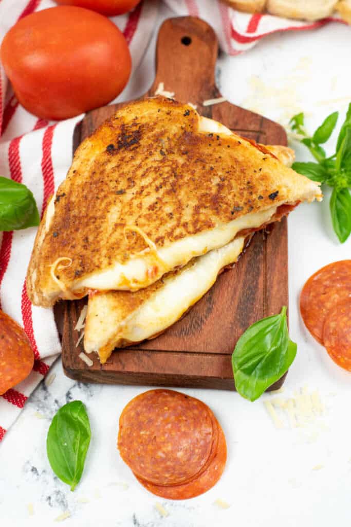 A grilled cheese sandwich but in half with white cheese melting out of it. It is on a small wooden chopping block surrounded by pepperoni and fresh basil leaves. 