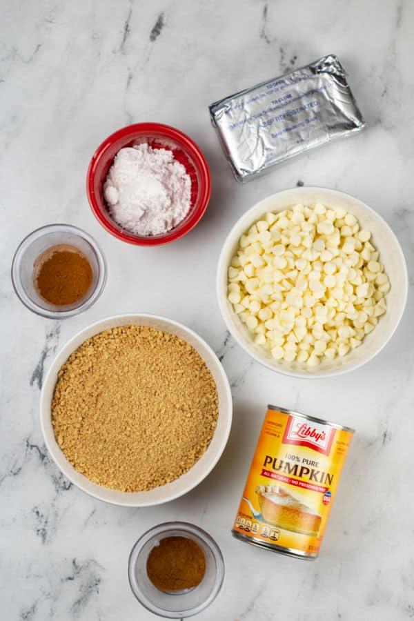 The ingredients for white chocolate truffles on a a white marble counter top. Cream cheese, powdered sugar, pumpkin pie spice, white chocolate chips, graham cracker crumbs, pumpkin puree and cinnamon. 