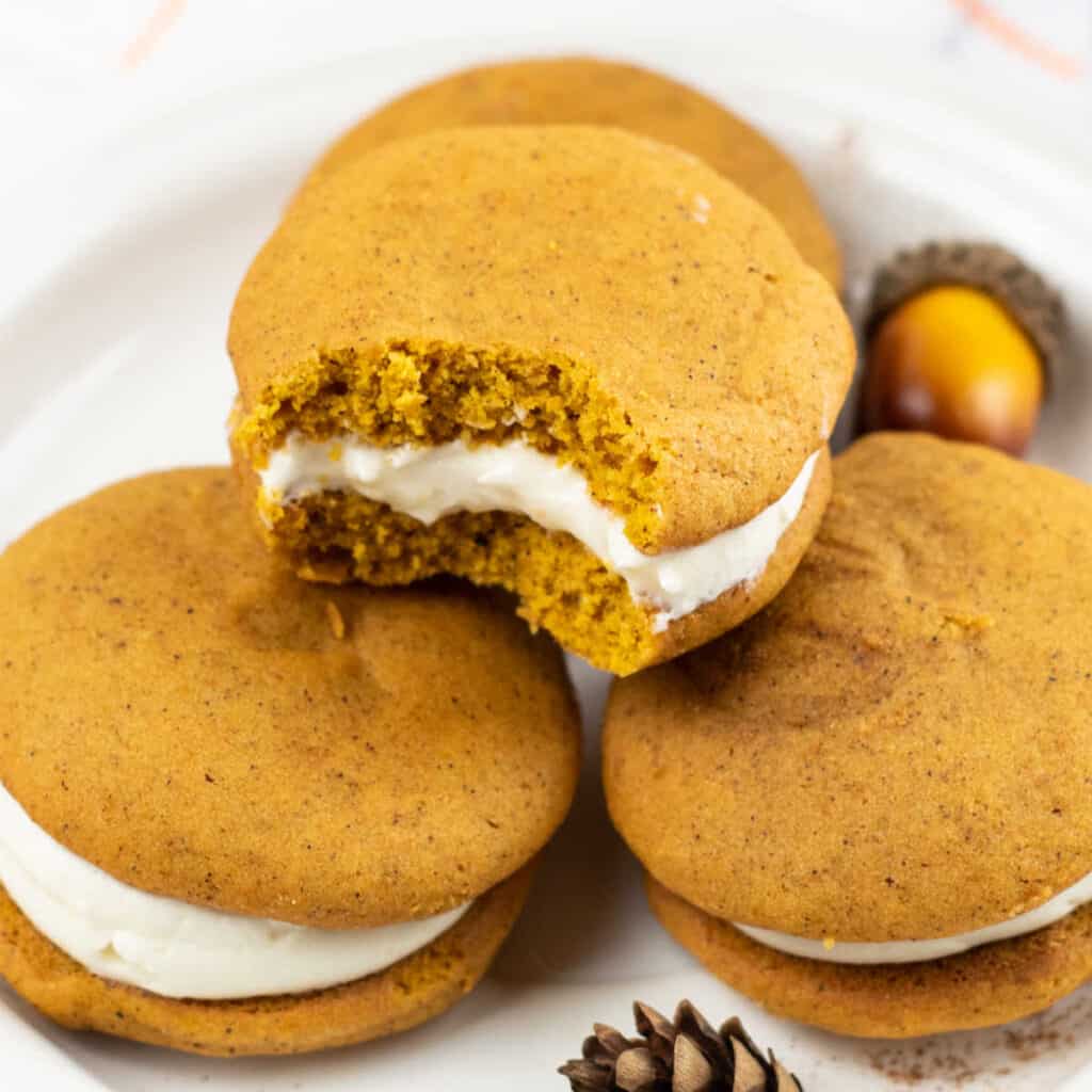 A pile of pumpkin whoopie pies, pumpkin cookies with a creamy filling. The one on the top has a bite taken out. 