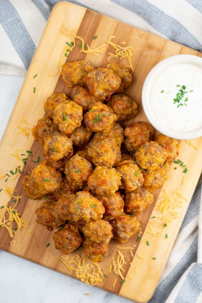 A pile of cheesy sausage balls on a wooden board with a bowl of dipping sauce next to them. 