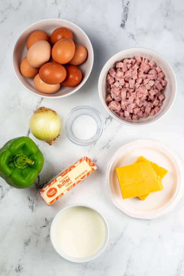The ingredients for a Denver omelet on a white marble counter top. Eggs, diced ham, cream, cheddar cheese, butter, green pepper and onion