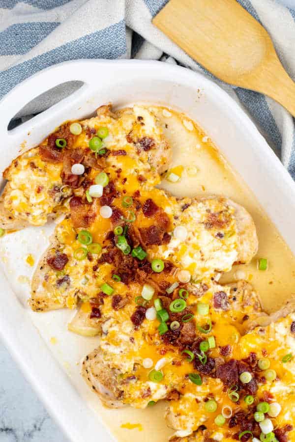 A white casserole dish with chicken breasts covered in melted cheese, bacon and green onions. 