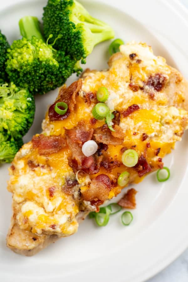 A white plate with a piece of chicken covered in cheese, bacon and green onions. On the side is some steamed broccoli. 