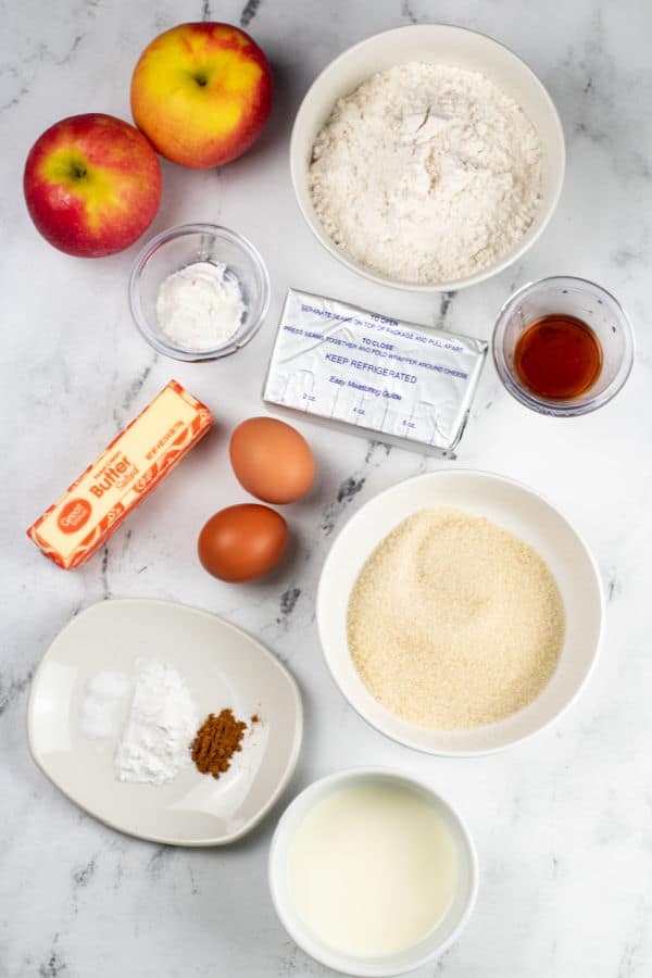 A white marble counter top with the ingredients for apple cream cheese coffee cake. Apples, flour, corn starch, vanilla, cream cheese, butter, eggs, sugar, milk, baking powder, salt and cinnamon. 
