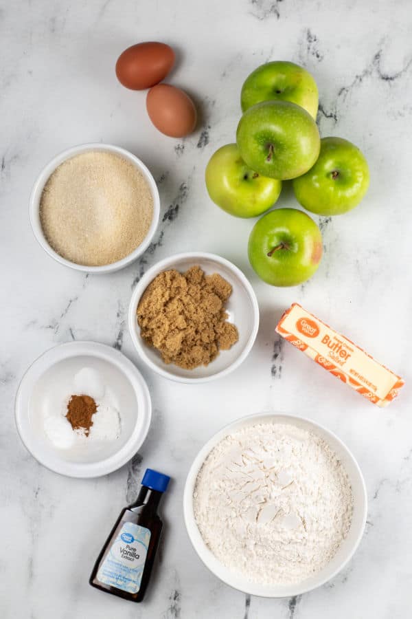 The ingredients for apple cinnamon muffins on a marble countertop. Eggs, granny smith apples, sugar, brown sugar, flour, butter, vanilla, salt, cinnamon, baking soda and baking powder. 