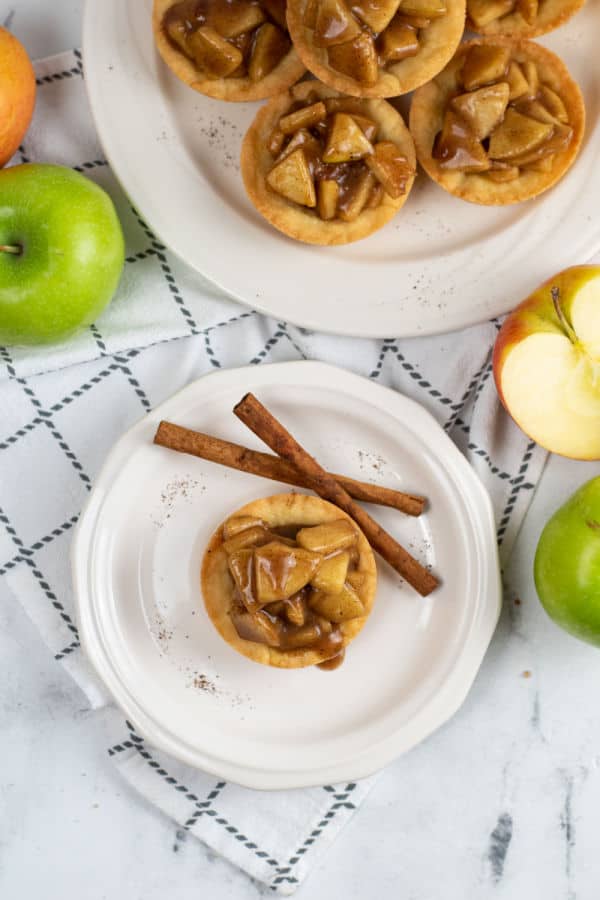 An overhead view of a table with a large plate of apple pie cookie cups and a smaller plate with just one. They are surrounded by cinnamon sticks and chopped apples. 