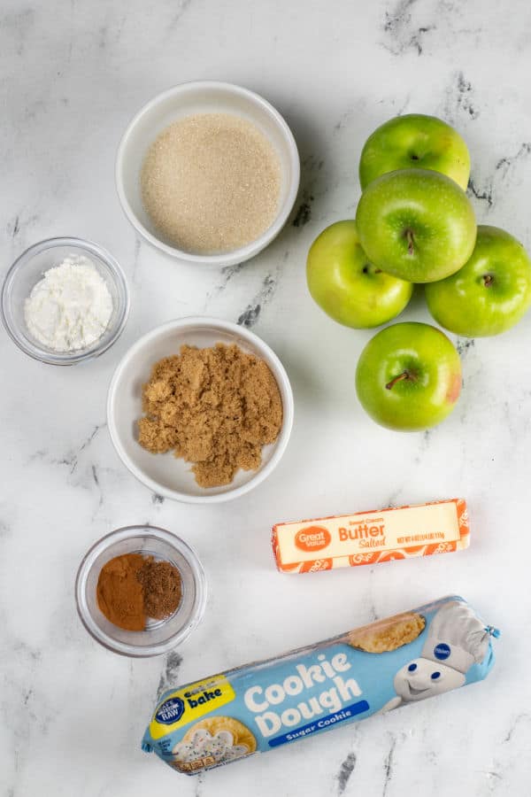 The ingredients for apple pie cookie cups on a white marble background. Sugar, apples, corn starch, brown sugar, butter, cinnamon, nutmeg and cookie dough. 