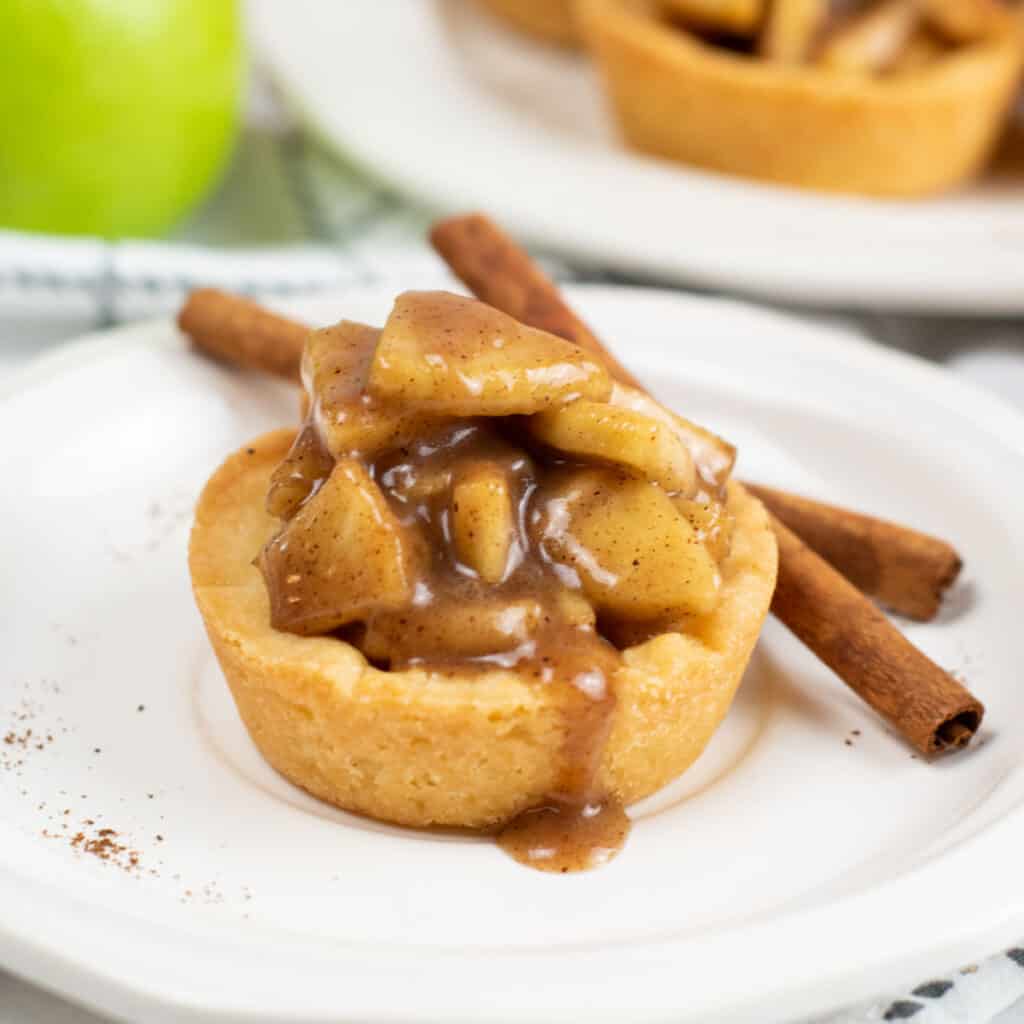 A close up of a apple pie cookie cup. A small pie with a cookie crust filled with apple pie filling, sitting on a white plate. 