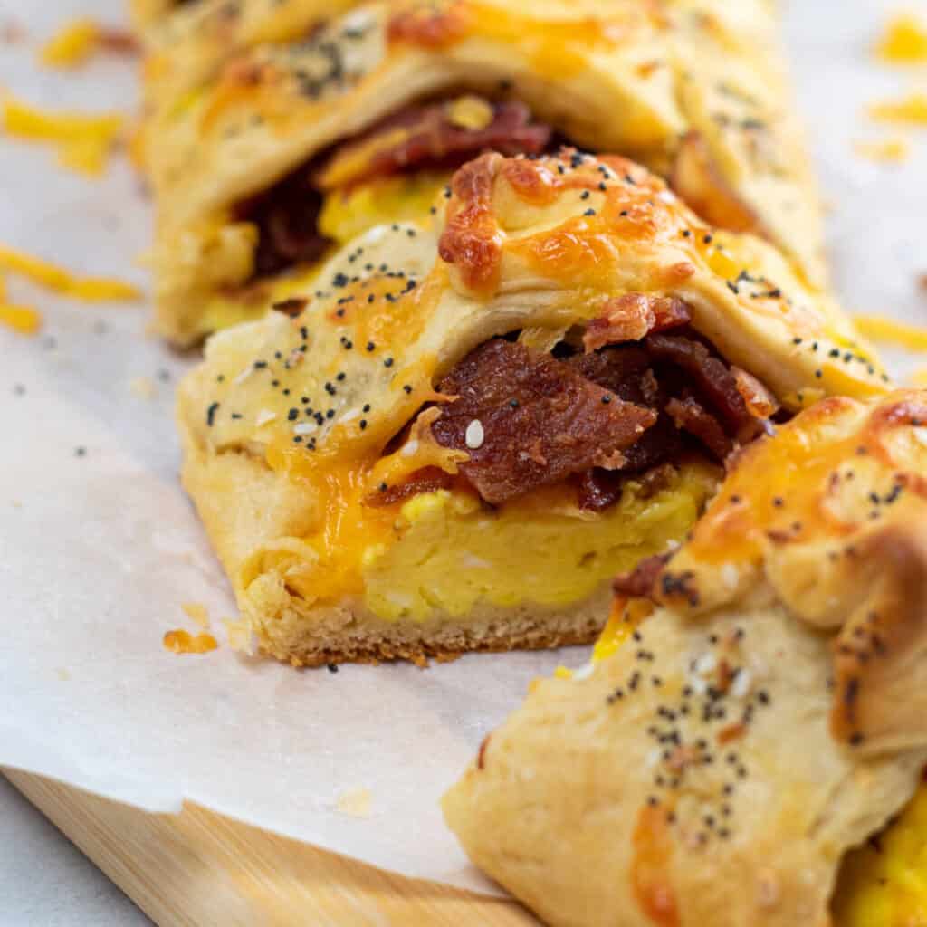 A close up of a slice of bacon egg & cheese breakfast braid. There is bacon and eggs wrapped in crescent roll and sprinkled with everything bagel seasoning
