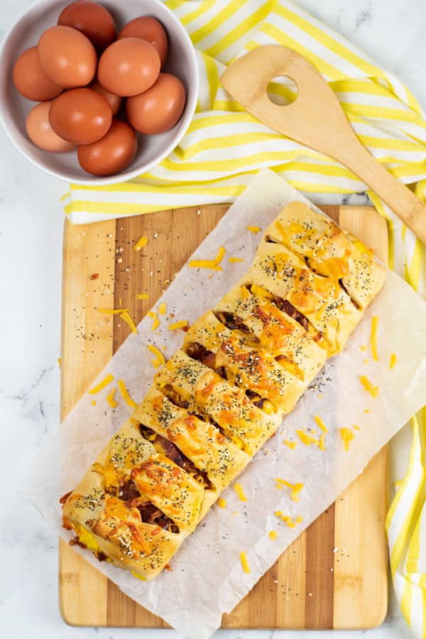 An overhead shot of a crescent roll breakfast braid on a wooden chopping board. Next to it sits a wooden spatula and a bowl full of eggs. 