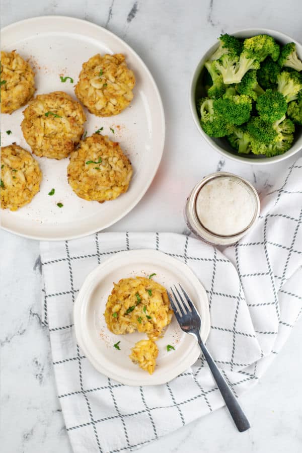 A marble counter top with a plate full of crab cakes, a bowl of steamed broccoli, a Mason jar full of beer and a plate with a crab cake and fork on it 