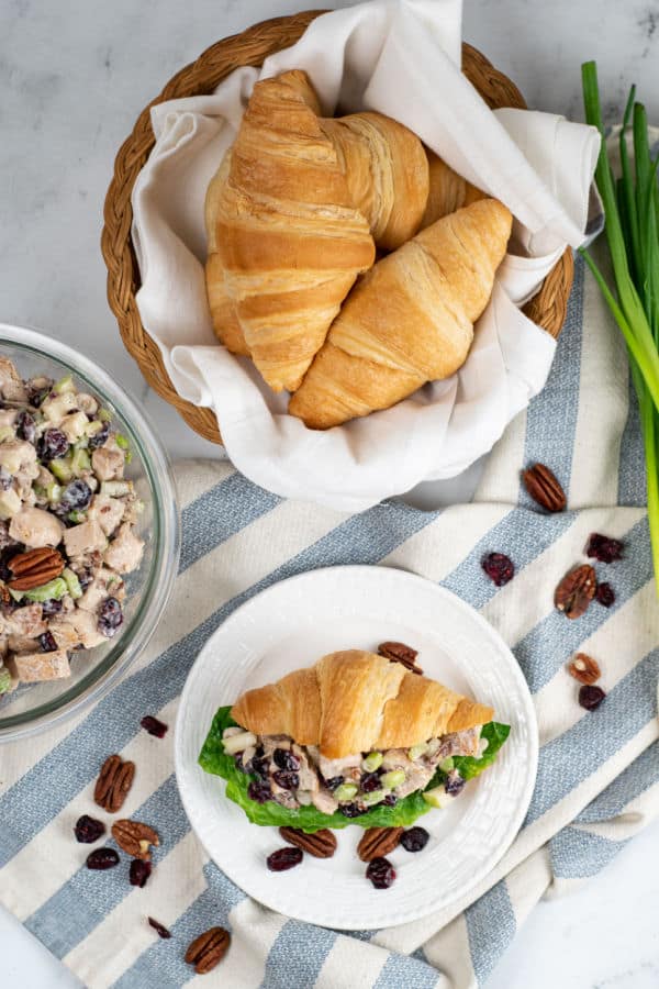 A table with a basket full of croissants, a clear glass bowl full of cranberry pecan chicken salad, and a white plate with a croissant and chicken salad sandwich garnished with pecans and dried cranberries.  