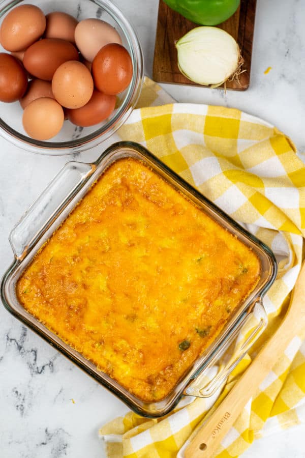 A square pan with cheese topped baked eggs next to the pan is a yellow and white linen, bowl of brown eggs and a chopping block with an onion and green pepper. 