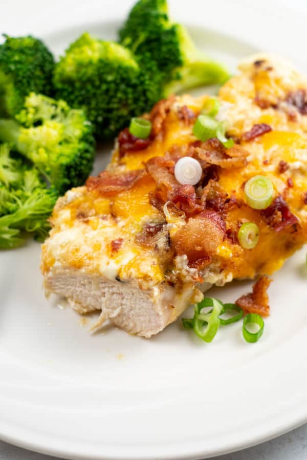 A piece of million dollar chicken on a plate with broccoli. The chicken is a juicy chicken breast smothered in melted cheeses, bacon pieces and green onion slices. 