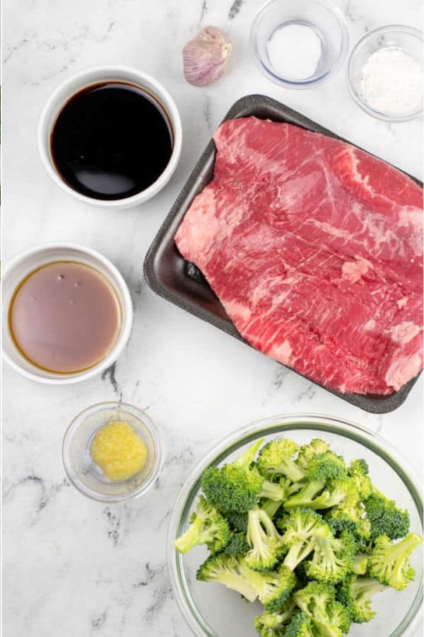 A white marble counter top with the ingredients for sheet pan beef and broccoli. Flank steak, broccoli florets, soy sauce, sesame oil, garlic, ginger, baking soda and corn starch. 