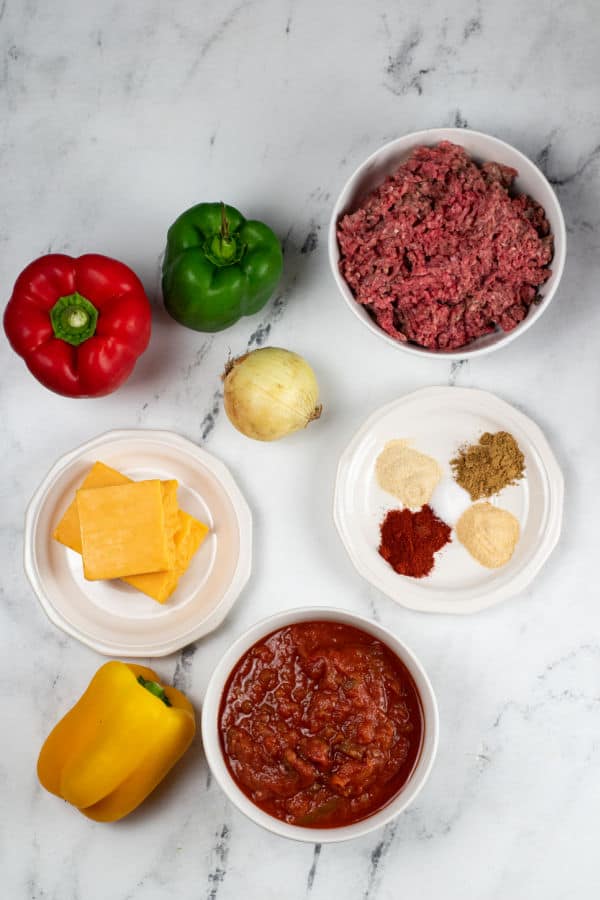 The ingredients for taco stuffed peppers on a white marble counter top. Ground beef bell peppers, onion, cheese, salsa, garlic, onion powder, smoked paprika, cumin and salt. 
