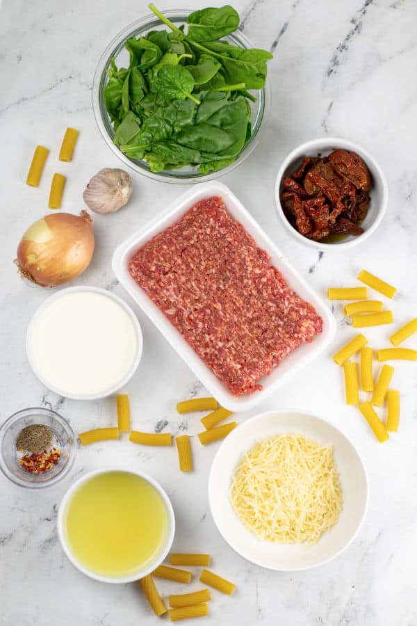 The ingredients for creamy Italian sausage pasta on a white marble countertop. Italian sausage, spinach, sun dried tomatoes, rigatoni, cream, chicken broth, pepper, red pepper flakes, onion and garlic. 