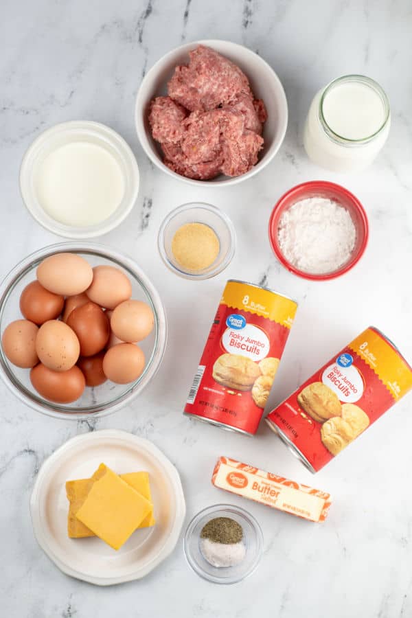 A white marble counter top with the ingredients for sausage biscuits and gravy casserole. Sausage, milk, cream, garlic powder, flour, eggs, biscuits, butter, salt and pepper and cheese. 