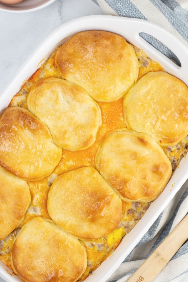 A white baking dish with a sausage biscuit and gravy casserole in it. The top is covered with golden brown biscuits. 