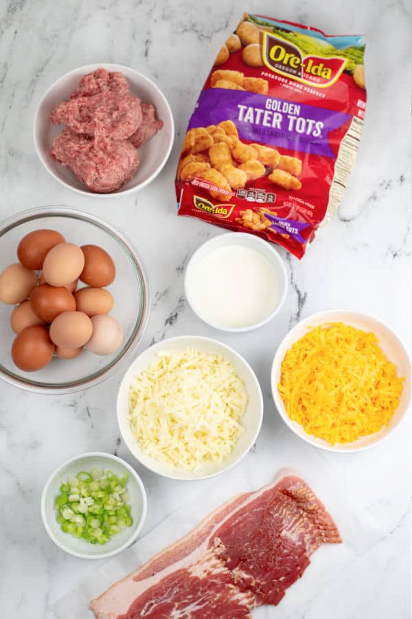 The ingredients for tater tot breakfast casserole on a white marble counter top. Tater tots, sausage, eggs, bacon, cream, shredded Monterey jack cheese, shredded cheddar cheese, and sliced green onion. 