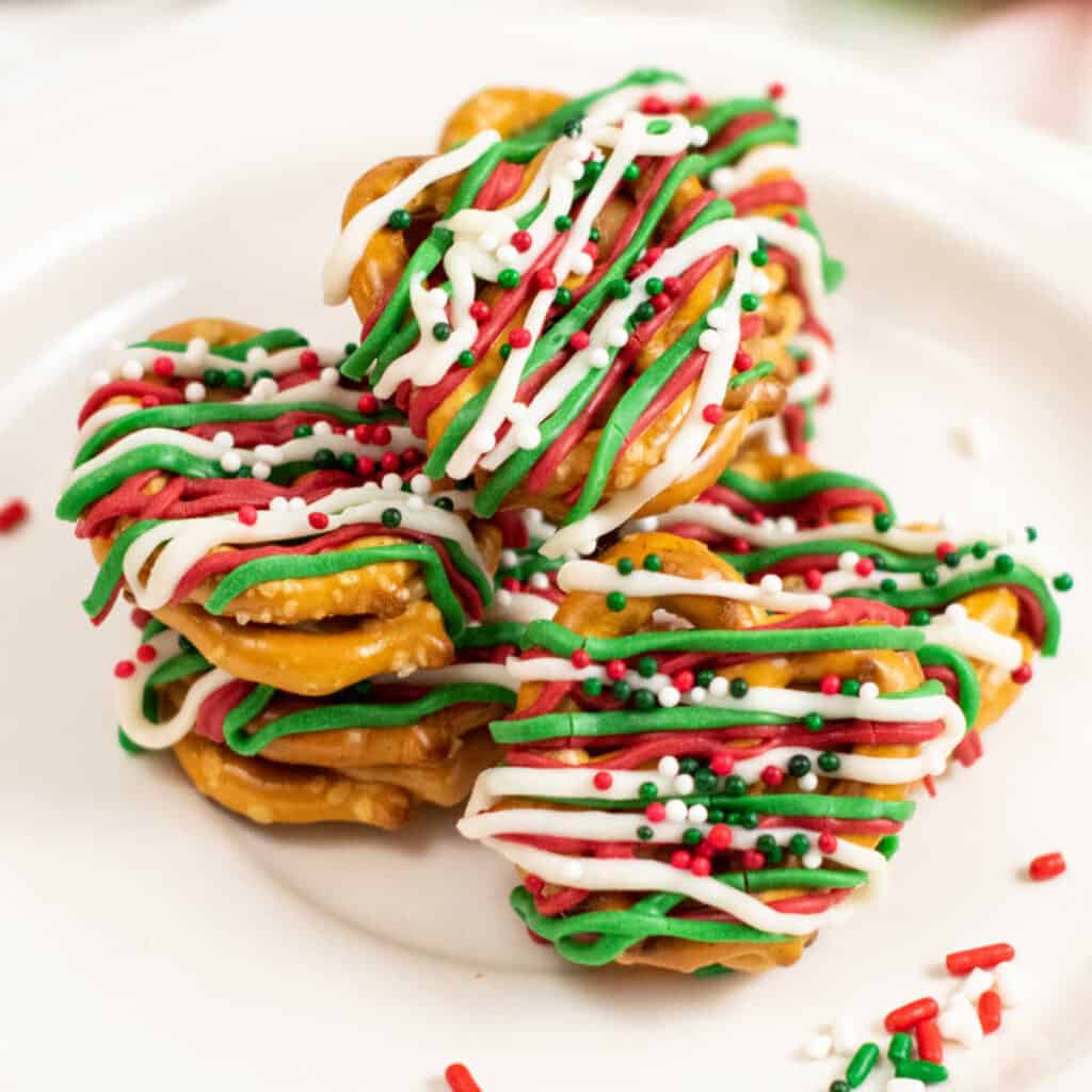 A pile of caramel filled pretzels drizzled with red, green and white chocolate and sprinkled with sprinkles. 