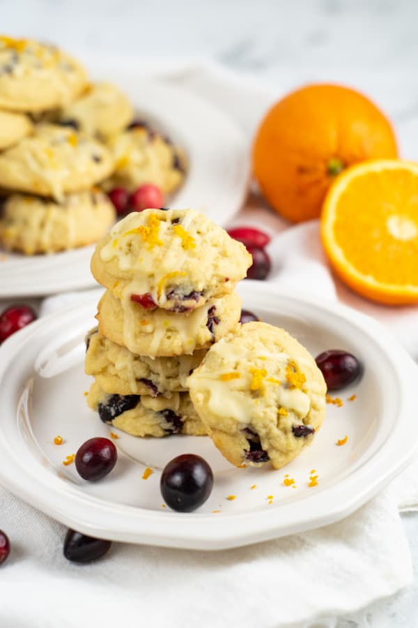 A stack of cranberry orange cookies on a white plate. In the background are oranges and a plate full of cookies. 